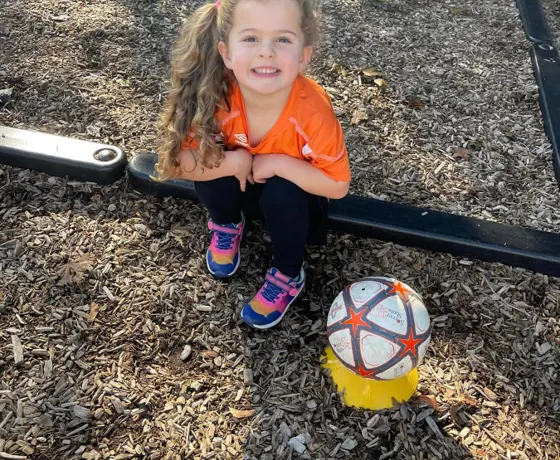 A smiling child sitting outside next to a soccer ball
